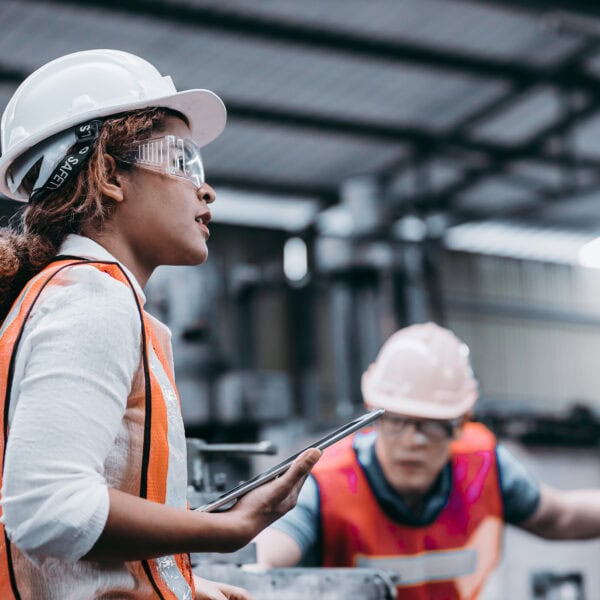 Female industrial engineer wearing a white helmet while standing in a heavy industrial factory behind she talking with workers, Various metal parts of the project